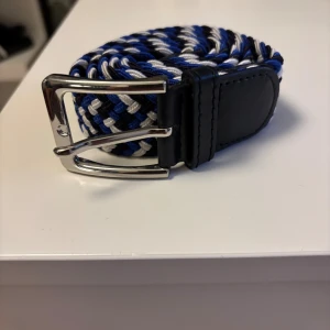 Randigt flätat bälte i blått och vitt - Snyggt flätat bälte med randigt mönster i blått, svart och vitt. Bältet har ett silvrigt spänne och detaljer i svart skinn. Perfekt för att ge outfiten lite extra edge och passar till både jeans och chinos.