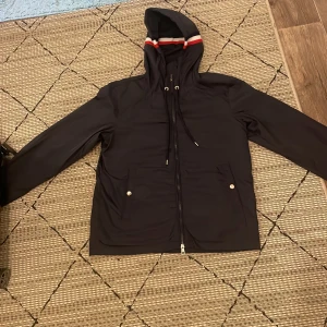  vindjacka från Moncler med huva - vindjacka från Moncler med huva och snygga röda och vita ränder på huvan. Jackan har dragkedja framtill, snörning vid huvan och Moncler-logga på bröstet. Knappar med Moncler-tryck och lätt material som passar perfekt för blåsiga dagar.