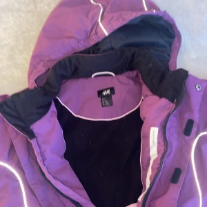 Lila vinterjacka med huva från H&M - Lila vinterjacka från H&M med svart och rosa detaljer, reflexränder och avtagbar huva. Jackan har dragkedja, muddar vid ärmslut och är fodrad med mjukt material på insidan. Perfekt för kalla dagar och har en sportig vibe.