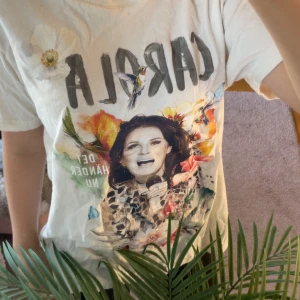 Vit turné-tshirt Carola 2024 - Vit t-shirt med färgglatt tryck framtill och texten 'CAROLA' samt blommor och fåglar. Baksidan har turnélistan 'DET HÄNDER NU TOUR 2024'. Klassisk passform och rund hals. Perfekt för dig som gillar musik och vill sticka ut med coolt motiv.