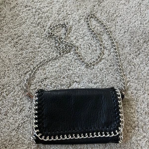 Svart axelväska med kedja från Tiamo - Cool svart axelväska från Tiamo i skinnimitation med silverfärgad kedja som axelrem. Väskan har dekorativa kedjedetaljer runt kanterna och stängs med magnetlås. Perfekt storlek för det viktigaste och ger en edgy vibe till din outfit. Ena magneten är lite trasig men funkar ändå (se bild 3). Den är lite rostig vid själva hakrn vid kedjans slut och början! (Se bild 4) Priset kan diskuteras! Kontakta mig ifall du behöver fler bilder eller bara har frågor!💗💗