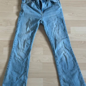 Blå bootcut jeans Bailey 158 - Snygga ljusblå bootcut jeans från Bailey i storlek 158. Jeansen har klassisk femficksdesign, resår i midjan och raka ben som slutar i en lätt utsvängd bootcut. Perfekta för dig som gillar en retro vibe och vill ha bekväma jeans i mjukt denimtyg.