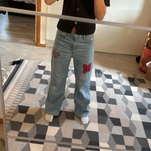Ljusblå jeans med patches och brodyr - Säljer ett par halvbaggy ljusblå jeans med bredare passform och coola patches på benen, bland annat ett stort rött M och broderade detaljer. Jeansen har klassisk femficksdesign och är i mjukt denimtyg. Perfekta för dig som vill sticka ut lite extra! Ena märket är lite bort tvättat och änden av benen är lite trasigt.