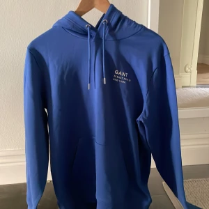 Blå hoodie från GANT med tryck - Helt ny GANT hoodie säljer pågrund av att den inte passade. Snyggt tryck på bröstet. Ny pris 1600 priset går  att förhandla ner vid snabb affär.