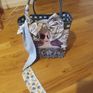 Svartvit shoppingväska med rosett och blommor - Unik shoppingväska i svartvitt rutmönster med stora handtag. Väskan är dekorerad med en stor mönstrad rosett, tygband i ljusblått och blommiga detaljer framtill. Perfekt för dig som gillar personliga accessoarer och vill sticka ut.