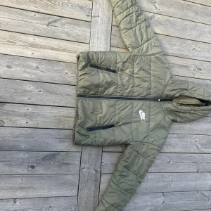 Nike Therma-Fit grön pufferjacka - Snygg grön pufferjacka från Nike med Therma-Fit-teknologi. Jackan har huva, två dragkedjefickor framtill och  Nike-logga på bröstet och ryggen. Perfekt för kyliga dagar och har en clean, sportig look med quiltad design och hel dragkedja.