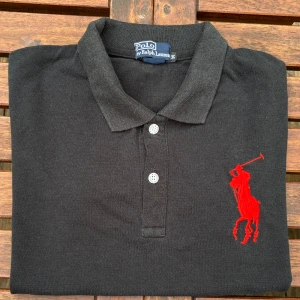 Svart pikétröja Polo Ralph Lauren - Svart pikétröja från Polo Ralph Lauren med klassisk krage, tre knappar och stor röd broderad logga på bröstet. På ena ärmen finns en röd siffra 3. Tröjan är i bomull och har korta ärmar. Perfekt för en clean och sportig look.