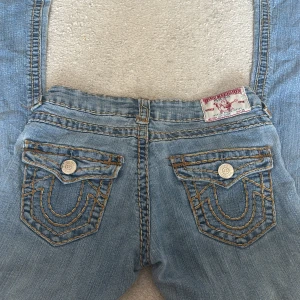 True Religion ljusblå jeans  - Säljer ett par ikoniska ljusblå jeans från True Religion med tydliga kontrastsömmar i gult och snygga ficklock bak med stora silverknappar. Klassisk femficksmodell och låg midja, tillverkade i mjuk bomullsdenim. 