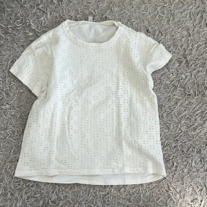 Vit broderad t-shirt i bomull - Fräsch vit t-shirt med korta ärmar och rund hals. T-shirten har ett broderat hålmönster över hela tyget som ger en cool och luftig vibe. Tillverkad i mjuk bomull, perfekt för varma dagar. Passformen är normal och enkel att styla till jeans eller shorts.