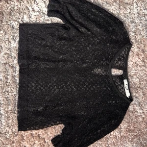 Svart spetsblus från NA-KD - Säljer en svart blus från NA-KD i genombruten spets med korta ärmar och rund halsringning. Blusen har en liten knapp i nacken och är perfekt för dig som gillar en stilren men ändå edgy look.