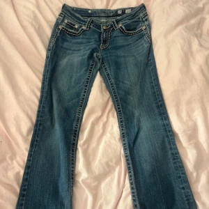 Blå bootcut jeans från Miss Me - Snygga blå jeans från Miss Me med bootcut passform och låg midja. Jeansen har dekorativa vita sömmar, detaljerade fickor med nitar och broderier samt klassisk femficksdesign. Materialet är slitstarkt denim och de har coola knappar med mönster. Midjemått: 36-38cm Innebenslängd: 80cm