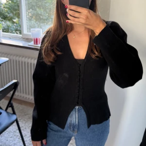 Svart tröja med knappar från Gina Tricot - Snygg svart tröja från Gina Tricot med v-ringning och dekorativa knappar framtill. Modellen är figurnära med lång ärm. Går att styla med ett linne under men även utan. Kan dressas upp och ned. 