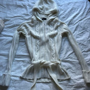 Vit stickad hoodie från Vero Moda - Säljer en vit stickad hoodie från Vero Moda med kabelstickat mönster, huva och knytskärp i midjan. Tröjan har långa ärmar, ribbade muddar och dragkedja framtill. Perfekt för lager-på-lager under kyliga dagar.