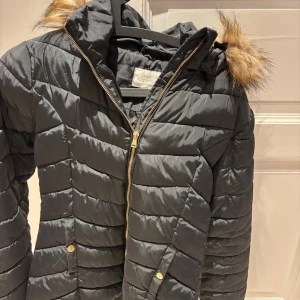 Svart pufferjacka med fuskpäls - Svart pufferjacka från KappAhl med guldig dragkedja och knappar. Jackan har huva med fuskpälsdetalj och quiltad design. Perfekt för kalla dagar och har en snygg, klassisk look som funkar till det mesta.