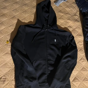 Svart hoodie från Polo Ralph Lauren - Svart hoodie från Polo Ralph Lauren med dragkedja och klassisk huva. Tröjan har en liten vit logga broderad på bröstet och praktiska fickor framtill. Tillverkad i mjuk bomull och har långa ärmar med muddar. Perfekt för en chill och stilren look.