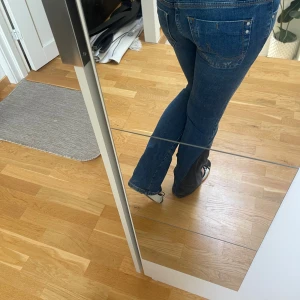 Blå bootcut jeans med slitningar - Snygga blå jeans med bootcut och slitna detaljer på låren och knäna. Jeansen har normal passform och klassiska bakfickor med nitar. Materialet är jeans och färgen är mörkblå med svarta inslag nertill på benen.