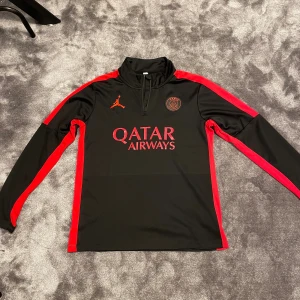 Psg tröja långärmad - Svart och röd långärmad PSG träningströja från Jordan med halv dragkedja, klubbmärke och Qatar Airways-tryck på bröstet. Tillverkad i funktionsmaterial som andas, med röda detaljer längs ärmar och sidor. Perfekt för fotbollsträning eller supporterliv.