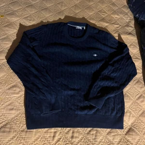 Mörkblå kabelstickad tröja från GANT - Säljer en mörkblå kabelstickad tröja från GANT med rund halsringning och broderad logga på bröstet. Tröjan har långa ärmar och ribbade muddar vid ärmslut och nederkant. Perfekt för dig som gillar klassisk och stilren look. Är storlek S men passar som M!