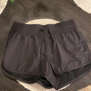 Svarta sportshorts från H&M - Svarta sportiga shorts från H&M med elastisk midja och snörning. Tillverkade i 100% polyester med mesh-detaljer längs sidorna för extra ventilation. Perfekta för träning eller löpning, med en loose passform som ger rörelsefrihet.