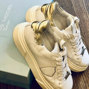 Philippe Model vita sneakers med guld - Snygga vita sneakers från Philippe Model Junior med metallicguld-detaljer på hälen och silvriga metalldetaljer på snörningen. Skorna har en chunky sula, rund tå och är tillverkade i skinn. Perfekta för dig som gillar stilrena men ändå unika sneakers. Skorna är knappt använda så är i nyskick.