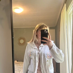 Vit kofta med broderade blommor - Superfin vit kofta från Cotton Philosophy med broderade blommor i pastellfärger och volangkrage. Koftan har knappar framtill, långärmade ärmar och romantiska detaljer längs kragen och ärmsluten. Perfekt för dig som gillar vintagevibbar och söta detaljer.💌