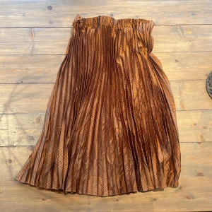 Brun plisserad långkjol Scotch & Soda - Säljer en brun plisserad långkjol från Scotch & Soda med tunna svarta diagonala ränder. Kjolen har ett elastiskt midjeband och är tillverkad i ett lätt, glansigt material som ger ett snyggt fall. Perfekt för dig som gillar coola detaljer och mönster.