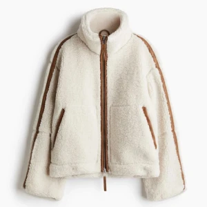 Vit teddyjacka med bruna detaljer -  fluffig vit teddyjacka med bruna detaljer. Jackan har hög krage, två stora fickor och en rak, 