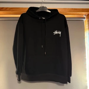 Svart hoodie från Stüssy - Svart hoodie från Stüssy med vit logga på bröstet och färgglatt grafiskt tryck på ryggen. Klassisk känguruficka framtill och justerbar huva med snören. Perfekt för en avslappnad streetwear-look.