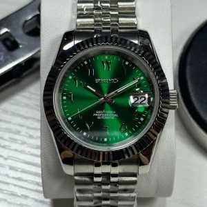 Grön automatisk klocka från Seiko - Snygg Seiko klocka med grön urtavla och arabiska siffror. Automatisk rörelse, datumvisning och robust silverfärgad länkarmband i metall. Urtavlan har en cool solbrun effekt och boetten är räfflad för extra stilpoäng.