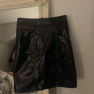 Svart skinnkjol -  svart kort skinnkjol,  har en rak passform och stängs med knapp