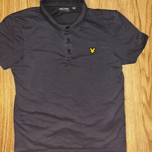 grå piké från Lyle & Scott - Snygg mörkgrå pikétröja från Lyle & Scott med klassisk krage och tre knappar framtill. Bröstet pryds av den ikoniska gula örnloggan. Tillverkad i mjuk bomull som känns skön mot huden. Perfekt för en clean och stilren look. 100% äkta lyle and scott. Perfekt kvalité. Skriv privat för mer frågor 