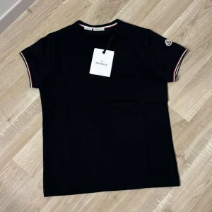 Svart Mon*ler t-shirt med logga. Svart - Svart t-shirt från Mon*ler i slim fit-modell. Klassisk design med korta ärmar och Mon*ler-logga på ena ärmen. Ribbade muddar med röd, vit och blå detalj vid ärmslut och halsringning. Tillverkad i mjuk bomull för en skön känsla.