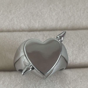 Silverring med hjärta och spjut - Cool och chunky silverfärgad ring med ett blankt hjärta i mitten och två spetsiga horn på sidorna. Ringen har en öppen baksida som gör den justerbar i storlek. Perfekt för dig som gillar edgy och unika accessoarer. Justerbar och rostfri 