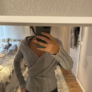 Grå omlottstickad tröja - Supersnygg grå stickad tröja med omlott-design och bred ribbad struktur över bröstet. Tröjan har lång ärm och en djup v-ringning som ger en modern och trendig look. Perfekt att styla med jeans eller kjol för en chill vibe.