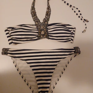 Randig bikini med smyckesdetaljer H&M - Snygg bikini från H&M med marinblå och vita ränder. Överdelen har halterneck med smyckesdetaljer och knyt i nacken, samt dekorativa pärlor framtill. Underdelen har matchande pärldetaljer på sidorna. Perfekt för stranden eller poolen! Änvända 1 gång nyskick.