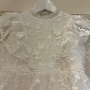Vit spetsklänning från Minimalina Bridal - Superfin vit kortklänning från Minimalina Bridal med broderade blommor och volangdetaljer. Klänningen har genomskinliga ärmar, dragkedja i ryggen och romantisk spets över hela plagget. Perfekt för dig som vill ha en dreamy och söt look.