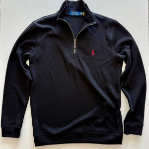 Svart half zip tröja från Polo Ralph Lauren - Svart långärmad tröja från Polo Ralph Lauren med half zip och klassisk krage. Tröjan har röd broderad logga på bröstet och är tillverkad i mjuk bomull. Perfekt för en stilren och avslappnad look.