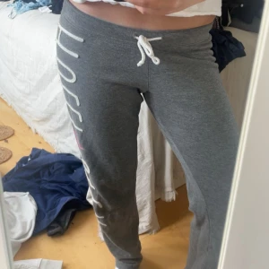 Grå mjukisbyxor från Hollister XS - Snygga grå mjukisbyxor från Hollister med vit snörning i midjan och stort tryck längs benet. Detalj med rosa paljetter på loggan. Sköna och mjuka, perfekta för chill eller träning. Passar dig som gillar casual och sportig stil.