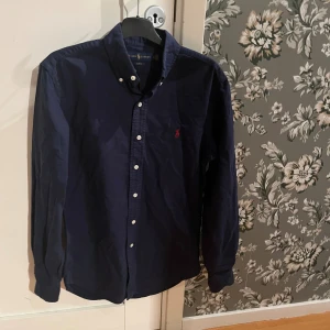 Mörkblå skjorta från Ralph Lauren - Säljer en mörkblå slim fit skjorta från Ralph Lauren med klassisk krage och vita knappar. Skjortan har lång ärm och den ikoniska röda loggan broderad på bröstet. Tillverkad i mjuk bomull, perfekt för en clean och stilren look.