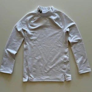 Vit långärmad träningströja Under Armour - Vit långärmad träningströja från Under Armour med hög krage och svart logga på halsen. Tröjan är tillverkad i ett stretchigt syntetmaterial som andas och passar perfekt för sport och träning. Enkel och stilren design med slitsar i sidorna.Det är en kompression tröja.