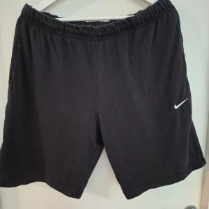 Svarta shorts från Nike - Svarta shorts från Nike med elastisk midja och klassisk vit swoosh-logga på ena benet. Modellen är loose fit och har en bakficka. Perfekta för träning eller chill dagar. Mjukt bomullsmaterial som känns skönt mot huden.