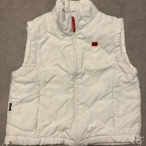 Vit quiltad dunväst från Diesel - Snygg vit dunväst från Diesel med quiltad design och hög krage. Västen har dragkedja framtill, diskret logga på bröstet och justerbar nederkant. Perfekt för lager-på-lager och ger en clean look till din outfit.