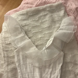 Vit volangbody från Amisu - Supersöt vit body från Amisu med v-ringning och stora volangdetaljer i transparent tyg längs halsringningen. Kroppsnära passform och ärmlös design, perfekt för att skapa en romantisk look. Materialet är mjukt och stretchigt för extra komfort.