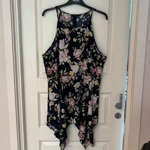 Blommig marinblå kortklänning  - Supersöt marinblå kortklänning med blommigt mönster i rosa, vitt och gult. Klänningen har smala axelband, asymmetrisk nederkant och dragkedja i ryggen. Perfekt för sommarens alla tillfällen!