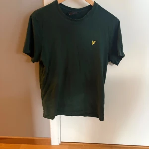 Grön t-shirt från Lyle & Scott - Snygg mörkgrön t-shirt från Lyle & Scott med klassisk rund hals och korta ärmar. På bröstet sitter den ikoniska gula fågel-loggan. T-shirten är gjord i mjuk bomull och har en normal passform som är chill och enkel att matcha.
