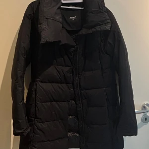 Svart medellång lättvikts vinterjacka från Everest - Säljer en svart medellång lättviktts vinterjacka från Everest med quiltad design och dragkedja framtill. Jackan har hög krage och långa ärmar, perfekt för kalla dagar. Stilren look och skön passform som håller dig varm hela vintern.