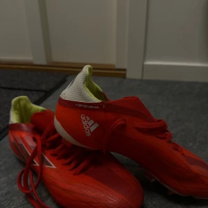 Röda Adidas fotbollsskor med dobbar - Säljer ett par röda Adidas fotbollsskor med vita och neongula detaljer. Skorna har snörning, låg profil och dobbar för bra grepp på planen. Tillverkade i syntetmaterial med sportig design och klassisk Adidas-logga på sidan.