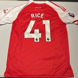 Arsenal Rice 41 matchtröja Adidas - Arsenal hemmatröja med Rice 41-tryck, officiell Premier League badge på ärmen. Röd kropp med vita ärmar och röda ränder, broderad Arsenal-logga och Adidas-märke. Tillverkad i lätt, ventilerande polyestermaterial med subtilt mönster i tyget. Replika, endast handtvätt gäller!