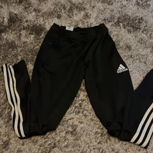 Svarta Adidas träningsbyxor S - Svarta träningsbyxor från Adidas med klassiska vita ränder längs benen och Adidas-logga på låret. Byxorna har smal passform, resår i midjan och är gjorda i ett mjukt och stretchigt polyestermaterial. Perfekta för sport eller chill.