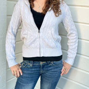 Vit stickad hoodie från Ralph Lauren - Vit kabelstickad hoodie från Ralph Lauren Sport i 100% bomull. Tröjan har dragkedja framtill, huva och ribbade muddar. Klassisk design med broderad logga på bröstet. Perfekt för en chill och stilren look.